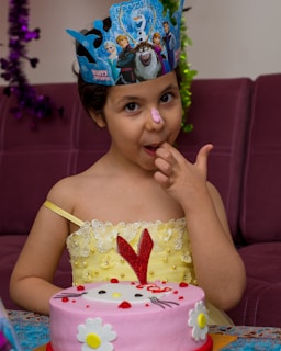 A young child wearing a decorative paper crown stands in front of a cake. The child has a smudge of pink frosting on their nose and is licking their finger. The cake is pink with a cartoon character design and flower decorations. The child wears a yellow dress adorned with floral patterns.