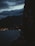 A silhouette of a person smoking against a backdrop of a dark, cloudy sky and a body of water with distant city lights twinkling. Mountains are visible in the background, creating a serene yet mysterious atmosphere.