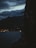 A silhouette of a person smoking against a backdrop of a dark, cloudy sky and a body of water with distant city lights twinkling. Mountains are visible in the background, creating a serene yet mysterious atmosphere.