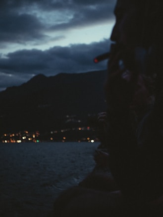 A silhouette of a person smoking against a backdrop of a dark, cloudy sky and a body of water with distant city lights twinkling. Mountains are visible in the background, creating a serene yet mysterious atmosphere.