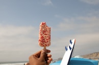 A hand holding an ice cream bar with a crunchy red and white coating. In the background, there are surfboards standing upright and a vast ocean under a partly cloudy sky.
