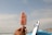 A hand holding an ice cream bar with a crunchy red and white coating. In the background, there are surfboards standing upright and a vast ocean under a partly cloudy sky.