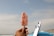 A hand holding an ice cream bar with a crunchy red and white coating. In the background, there are surfboards standing upright and a vast ocean under a partly cloudy sky.