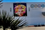 A colorful mural of a woman's face wearing a headdress made of vibrant flowers is displayed on a white tiled wall of a building. The building also features a sign for a bistro named 'Lulu'. In the foreground, spiky plant leaves are slightly silhouetted against the lighter wall.