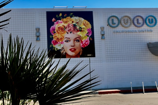 A colorful mural of a woman's face wearing a headdress made of vibrant flowers is displayed on a white tiled wall of a building. The building also features a sign for a bistro named 'Lulu'. In the foreground, spiky plant leaves are slightly silhouetted against the lighter wall.