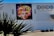 A colorful mural of a woman's face wearing a headdress made of vibrant flowers is displayed on a white tiled wall of a building. The building also features a sign for a bistro named 'Lulu'. In the foreground, spiky plant leaves are slightly silhouetted against the lighter wall.