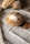 A loaf of round artisanal bread is placed on a wooden cutting board, surrounded by two other types of bread on a linen cloth. The bread appears freshly baked, with a crisp and lightly floured crust. The background includes a rustic, textured fabric and a lacy edge.