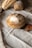 A loaf of round artisanal bread is placed on a wooden cutting board, surrounded by two other types of bread on a linen cloth. The bread appears freshly baked, with a crisp and lightly floured crust. The background includes a rustic, textured fabric and a lacy edge.