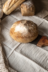A loaf of round artisanal bread is placed on a wooden cutting board, surrounded by two other types of bread on a linen cloth. The bread appears freshly baked, with a crisp and lightly floured crust. The background includes a rustic, textured fabric and a lacy edge.