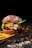 A close-up of a gourmet burger featuring a dark bun topped with flour, layers of lettuce, melted cheese, and a thick beef patty, set against a dark background with scattered spices and powder.