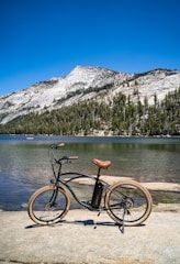 A black bicycle with brown seat and tires is parked on a stone surface next to a serene lake. In the background, a mountain range covered with green pine trees is visible under a clear, vibrant blue sky.