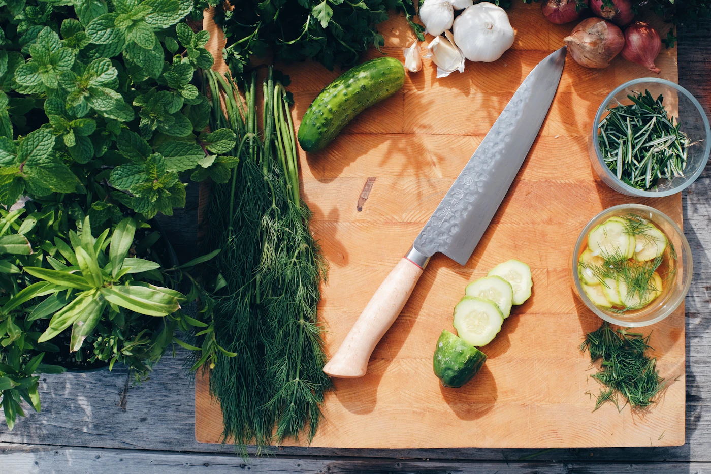 An overhead prep board with fresh mint, parsley and dill, garlic, shallots and chopped rosemary beside a chef's knife