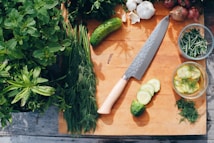A wooden cutting board holds a large, sharp knife and slices of cucumber. Surrounding the board are fresh herbs including dill, mint, and basil. There are garlic bulbs, shallots, and a whole cucumber. Bowls contain rosemary and a cucumber salad with dill.