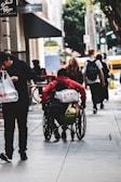 A person in a red hoodie sitting in a wheelchair moves along an urban sidewalk with other pedestrians. The person carries multiple bags attached to the wheelchair. A man in dark clothing is walking beside them holding a plastic bag. Other people can be seen walking in the distance, and there are trees and buildings in the background.