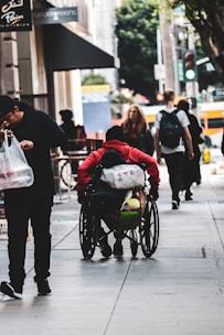 A person in a red hoodie sitting in a wheelchair moves along an urban sidewalk with other pedestrians. The person carries multiple bags attached to the wheelchair. A man in dark clothing is walking beside them holding a plastic bag. Other people can be seen walking in the distance, and there are trees and buildings in the background.