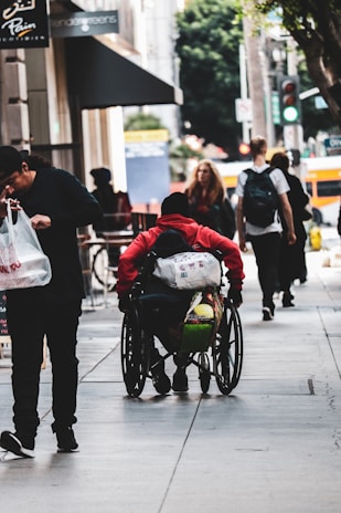 A person in a red hoodie sitting in a wheelchair moves along an urban sidewalk with other pedestrians. The person carries multiple bags attached to the wheelchair. A man in dark clothing is walking beside them holding a plastic bag. Other people can be seen walking in the distance, and there are trees and buildings in the background.