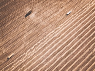 Aerial view of agricultural fields with distinct parallel lines indicating harvested areas. Two vehicles are present, possibly farm equipment, casting shadows on the textured landscape.