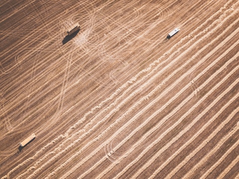 Aerial view of agricultural fields with distinct parallel lines indicating harvested areas. Two vehicles are present, possibly farm equipment, casting shadows on the textured landscape.