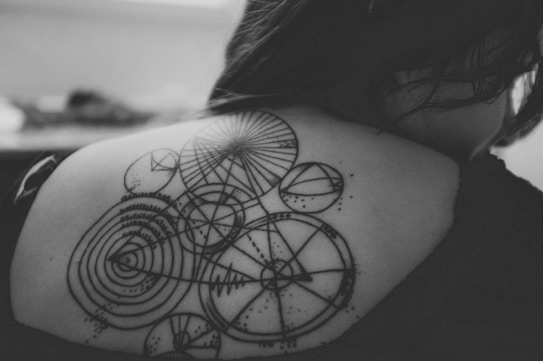 An intricate tattoo featuring geometric patterns and circles is displayed on the back of a person. The design consists of various circular shapes interconnected with lines and dots, creating an elaborate symmetrical pattern.