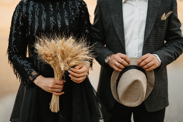 A couple is dressed in formal attire, with one person holding a bunch of dried wheat and the other holding a hat. The woman's dress is black and decorated with sequins, while the man wears a checkered blazer and a white shirt.