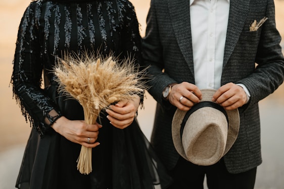 A couple is dressed in formal attire, with one person holding a bunch of dried wheat and the other holding a hat. The woman's dress is black and decorated with sequins, while the man wears a checkered blazer and a white shirt.