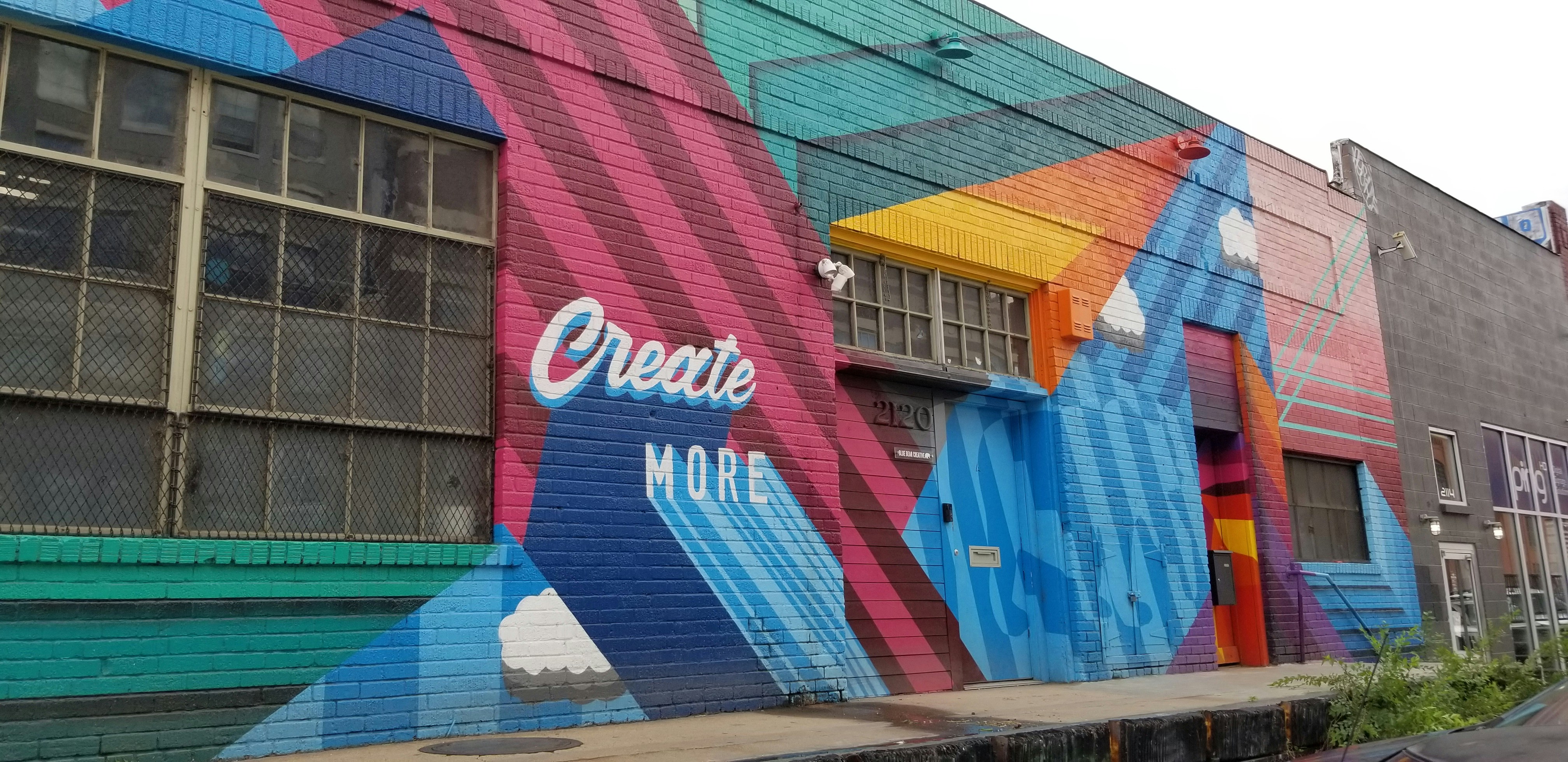 A colorful mural painted on the side of a building, featuring geometric shapes and patterns. The words 'Create More' are prominently displayed in white against a vibrant background of red, blue, teal, and orange. The artwork includes stylized clouds integrated into the design.