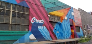 A colorful mural painted on the side of a building, featuring geometric shapes and patterns. The words 'Create More' are prominently displayed in white against a vibrant background of red, blue, teal, and orange. The artwork includes stylized clouds integrated into the design.