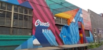 A colorful mural painted on the side of a building, featuring geometric shapes and patterns. The words 'Create More' are prominently displayed in white against a vibrant background of red, blue, teal, and orange. The artwork includes stylized clouds integrated into the design.