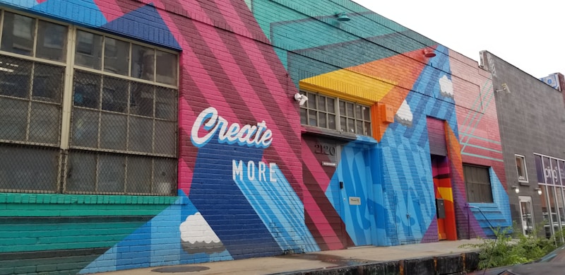 A colorful mural painted on the side of a building, featuring geometric shapes and patterns. The words 'Create More' are prominently displayed in white against a vibrant background of red, blue, teal, and orange. The artwork includes stylized clouds integrated into the design.