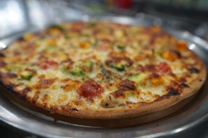 A freshly baked pizza with a golden-brown, crispy crust, topped with melted cheese, chunks of tomato, and green bell peppers. The pizza sits on a metallic tray with a blurred background, giving a warm and inviting appearance.