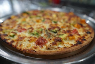 A freshly baked pizza with a golden-brown, crispy crust, topped with melted cheese, chunks of tomato, and green bell peppers. The pizza sits on a metallic tray with a blurred background, giving a warm and inviting appearance.