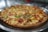 A freshly baked pizza with a golden-brown, crispy crust, topped with melted cheese, chunks of tomato, and green bell peppers. The pizza sits on a metallic tray with a blurred background, giving a warm and inviting appearance.