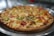 A freshly baked pizza with a golden-brown, crispy crust, topped with melted cheese, chunks of tomato, and green bell peppers. The pizza sits on a metallic tray with a blurred background, giving a warm and inviting appearance.