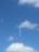 A tall radio or telecommunications tower extends skyward, set against a backdrop of a bright blue sky with scattered white clouds. The tower is thin and composed of sections that are alternately painted red and white. Support cables are visible, anchoring the structure.