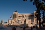 A grand, neoclassical building with ornate sculptures and columns stands majestically under a clear blue sky. Italian flags are prominently displayed, and a bus with the words 'ROMA CRISTIANA' can be seen in the foreground. Several people and vehicles are present near the large structure.