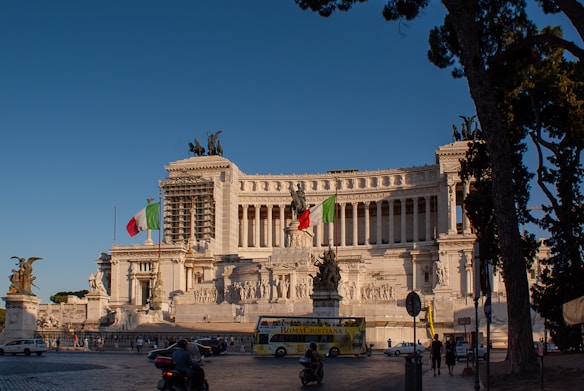 A grand, neoclassical building with ornate sculptures and columns stands majestically under a clear blue sky. Italian flags are prominently displayed, and a bus with the words 'ROMA CRISTIANA' can be seen in the foreground. Several people and vehicles are present near the large structure.