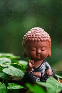 A small Buddha figurine is sitting in a meditative pose surrounded by lush green foliage. The figurine has a serene expression, and rain droplets appear to be falling around it, creating a tranquil atmosphere.