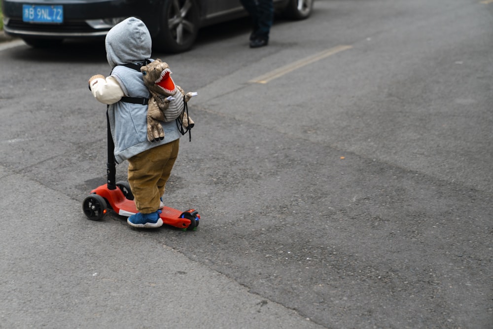 child in brown jacket riding red kick scooter on gray asphalt road during daytime