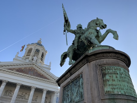 A bronze statue of a mounted rider holding a flag is prominently displayed against a clear blue sky. Behind the statue, a neoclassical building with columns and a clock tower is visible. The building is adorned with detailed architectural elements, and a flag can be seen flying near the top.