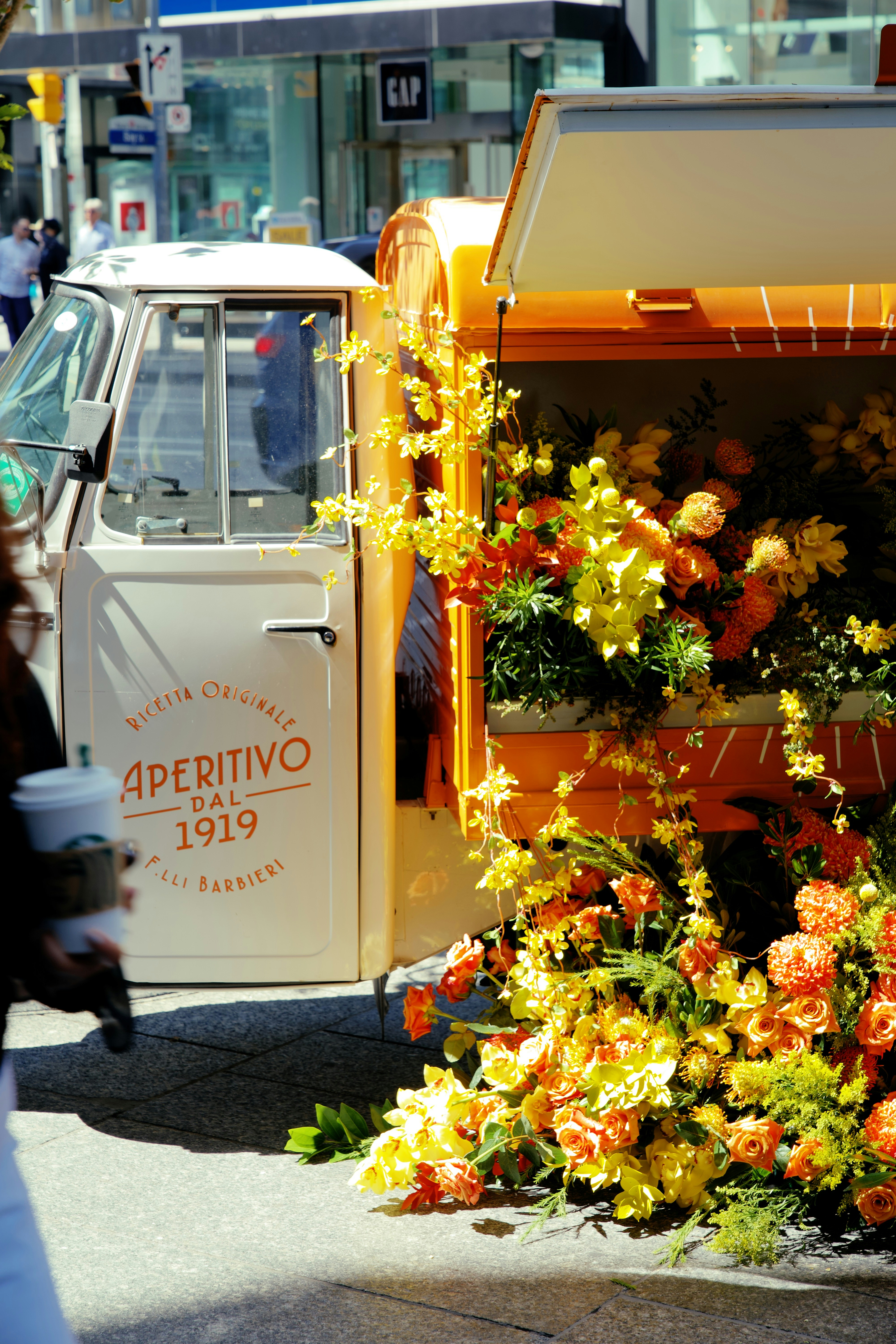 Street-side vintage delivery truck brims with a lush arrangement of orange and yellow flowers. The vibrant bouquet spills color onto the curb, creating a warm focal contrast against the urban backdrop.