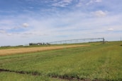 A vast agricultural field stretches out under a blue sky with a few scattered clouds. The landscape includes a grassy foreground, a section of golden wheat, and a green crop area. In the middle ground, a center pivot irrigation system extends horizontally across the farmland. The horizon is lined with trees and a small red-roofed structure is visible in the distance.