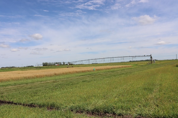 A vast agricultural field stretches out under a blue sky with a few scattered clouds. The landscape includes a grassy foreground, a section of golden wheat, and a green crop area. In the middle ground, a center pivot irrigation system extends horizontally across the farmland. The horizon is lined with trees and a small red-roofed structure is visible in the distance.