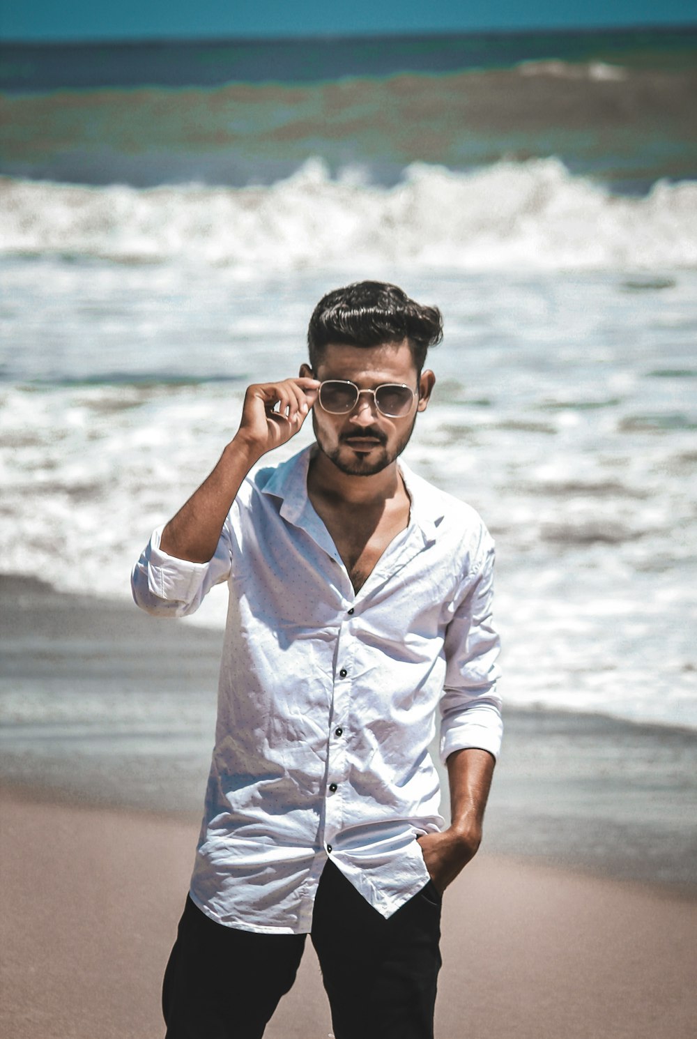 uomo in camicia bianca che indossa occhiali da sole neri in piedi sulla spiaggia durante il giorno