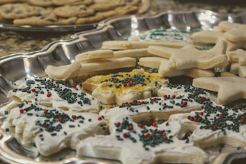 A silver tray holds a variety of sugar cookies, some frosted with white icing and decorated with red and green sprinkles. The cookies come in different shapes, such as stars and trees. In the background, there is another tray of plain cookies.
