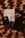 A close-up of luxury cosmetic items on a woven textured surface. The focus is on a black bottle with a logo, accompanied by a white box and another dark item, possibly a wallet or case. The scene is arranged artistically with dried floral elements adding an aesthetic touch.