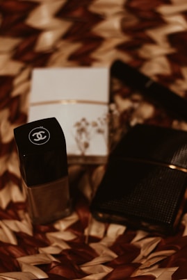 A close-up of luxury cosmetic items on a woven textured surface. The focus is on a black bottle with a logo, accompanied by a white box and another dark item, possibly a wallet or case. The scene is arranged artistically with dried floral elements adding an aesthetic touch.