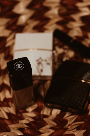 A close-up of luxury cosmetic items on a woven textured surface. The focus is on a black bottle with a logo, accompanied by a white box and another dark item, possibly a wallet or case. The scene is arranged artistically with dried floral elements adding an aesthetic touch.
