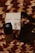 A close-up of luxury cosmetic items on a woven textured surface. The focus is on a black bottle with a logo, accompanied by a white box and another dark item, possibly a wallet or case. The scene is arranged artistically with dried floral elements adding an aesthetic touch.