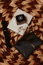 An arrangement of Chanel cosmetic products on a woven textured background. There is a black cylindrical tube, likely a lipstick or mascara, alongside a small black square container with the Chanel logo. A couple of white, textured, square items and some delicate dried flowers are also present.