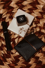 An arrangement of Chanel cosmetic products on a woven textured background. There is a black cylindrical tube, likely a lipstick or mascara, alongside a small black square container with the Chanel logo. A couple of white, textured, square items and some delicate dried flowers are also present.